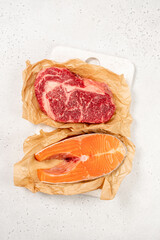 Cuts of beef and salmon are placed on parchment paper on a cutting board. They are ready for cooking in a kitchen setting, showing fresh ingredients for meal prep.
