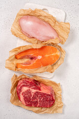 The scene shows three types of raw meat laid out on brown paper. There is chicken breast, beef steak, and salmon fillet ready for cooking in a kitchen setting.