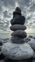Close-up of a tall stack of smooth stones balanced on a rocky beach under a cloudy sky. Symbol of balance and meditation.