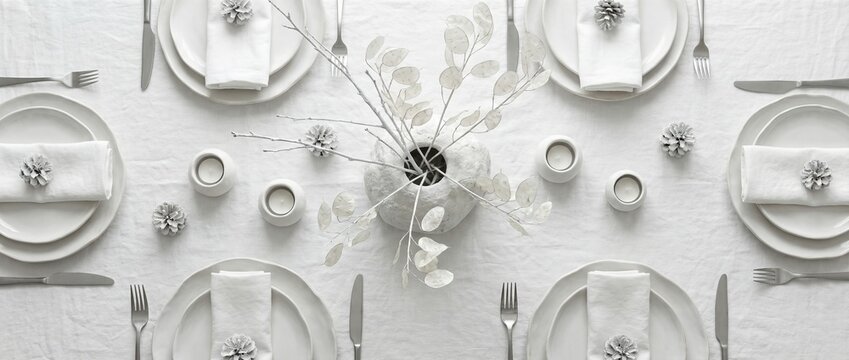 Elegant minimalist white table setting viewed from above. Neutral toned dinner arrangement with eucalyptus branches pine cones and simple dishware. Festive winter holiday dining concept for home - Powered by Adobe