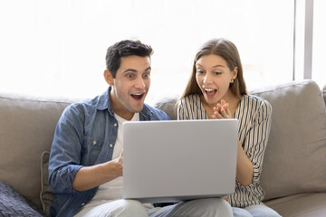 Couple sits on sofa, emotionally reacting to surprising news on laptop, celebrating unexpected success, got breakthrough announcements, winning prize, getting accepted to bank loan, relish lottery win