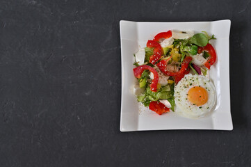 fresh salad with an egg on top, served on a white plate, captured against a dark surface. contrast in food photography. healthy salad with sliced eggs, sweet red pepper and lettuce.