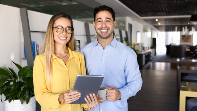 Two cheerful business partners posing in office workspace for portrait, holding tablet, looking at camera, smiling, enjoying leisure, coffee break, cooperation, teamwork. Banner shot
