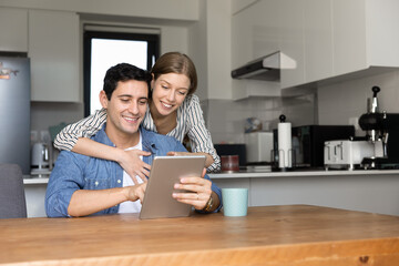 Young loving couple using modern digital tablet in cozy kitchen, buying goods through online supermarket platform or application, making order, discussing new app, spend pastime together on internet