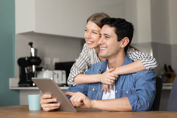 Cheerful young 30s couple in love using tablet device in cozy kitchen at home, booking travel together, enjoying online shopping, planning future. Modern lifestyle, digital connection, relationships