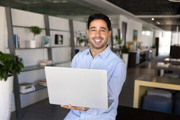Happy young Latin businessman holding laptop, posing in office co-working space, looking at camera, smiling. Business portrait of Hispanic professional, entrepreneur using computer