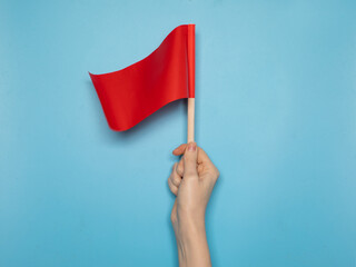 Hand holding a small red flag against a blue background. Symbol of alert, warning, danger sign or protest. Minimalist conceptual image. © Sergey Chumakov