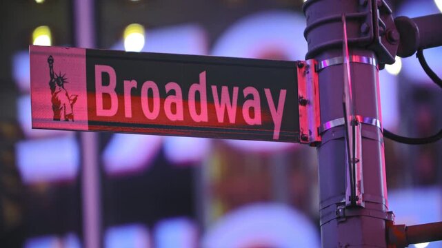 A vibrant, close-up shot of the iconic Broadway street sign in Manhattan, New York City, illuminated against a blurred background of city lights and billboards at night. 
