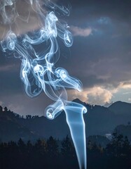 Wispy column of smoke against a cloudy, forested mountain backdrop