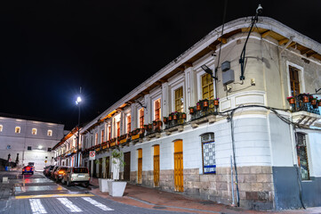 Colonial architecture lines San Francisco Square in historic center of Quito, Ecuador. White building features balconies with flower pots illuminated by streetlights under night sky
