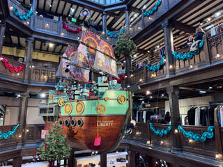 London, UK, A festive &ldquo;Good Ship Liberty&rdquo; installation loaded with wrapped gifts, suspended in the wooden-balconied atrium of Liberty London, decorated with garlands and watched by shoppers.