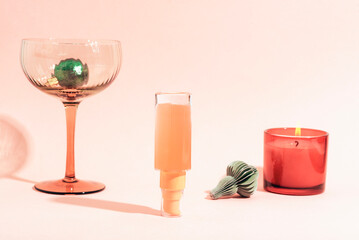 Cosmetic serum bottle standing upside down, candle, wineglass and Christmas ornaments on pink background. Holiday-themed beauty products, mockup