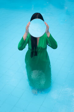Reflections in blue water: A portrait of disappearance. An anonymous woman in green dress with round mirror in swimming pool