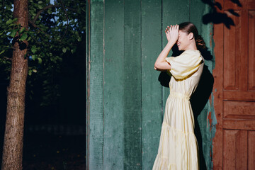 Woman in a pale yellow dress stands outdoors beside a green wooden doorway, shielding her eyes with a hand, creating a dramatic contrast of light and shadow.