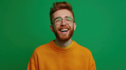Fototapeta premium A man with red beard and glasses laughs while wearing an orange sweater. He is inside and the background is green. This scene shows a casual moment in a bright setting.