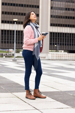 Mature woman standing on tiled plaza wearing pink sweater while holding coffee cup and smartphone