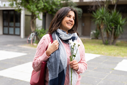 Woman holding daisies and smiling in courtyard by bicycle wearing blue scarf carrying red bag