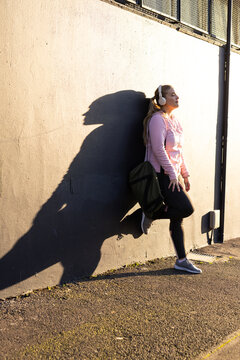 Woman leaning on wall at walkway, wearing pink hoodie, headphones, carrying green bag, copy space