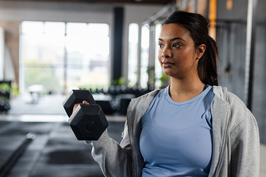 Asian female wearing tank top lifting hexagonal dumbbell at shoulder level in gym copy space