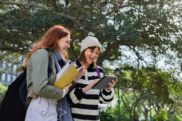 Diverse female students standing on campus path with notebooks, checking tablet, carrying backpacks