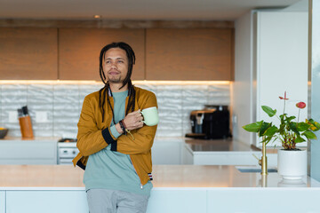 African American man wearing bomber jacket holding mug standing in home kitchen by coffee machine