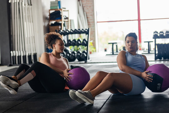 African American female friends sitting on gym mat twisting with medicine balls by dumbbell rack