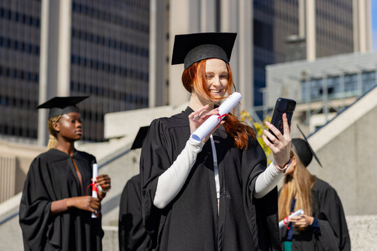 Diverse classmates posing on campus steps wearing mortarboards and holding diplomas and smartphones