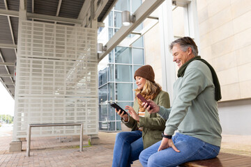Senior couple sitting on bench under metal canopy at transit platform checking devices, copy space