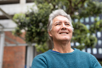 Senior man smiling, gazing upward in urban courtyard near modern building wearing teal sweater