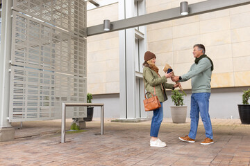 Senior couple exchanging paper-wrapped bouquet by screen panel on plaza under canopy, copy space