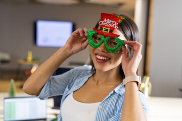 Smiling Indian woman adjusting green glitter glasses topped with red Christmas hat in office