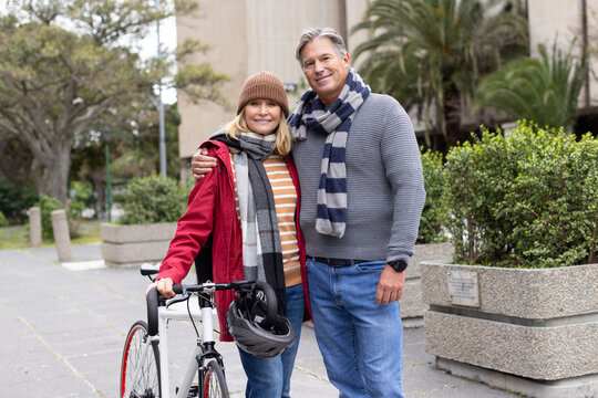 Senior couple posing on urban walkway with road bicycle and hanging helmet wearing scarf and beanie