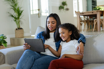 Mother and daughter sitting on sofa at home using tablet pointing at screen with potted plants