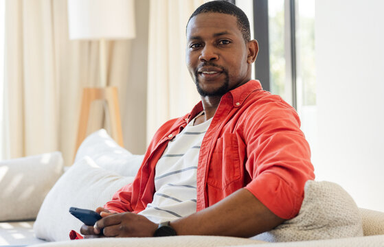 African American man sitting on sofa near floor lamp in living room holding smartphone, copy space