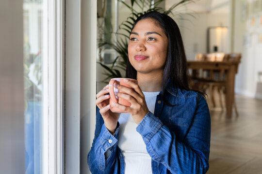 Woman standing inside home dining area near large window holding ceramic mug and looking outside