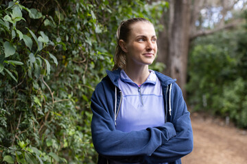 Woman standing on narrow dirt trail between hedge and trees, wearing athletic top and hoodie
