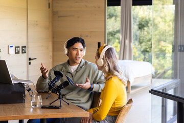 Diverse friends recording podcast at wooden table in home studio with microphone, headphones