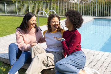Diverse female friends sitting on wooden pool deck beside backyard swimming pool and metal ladder