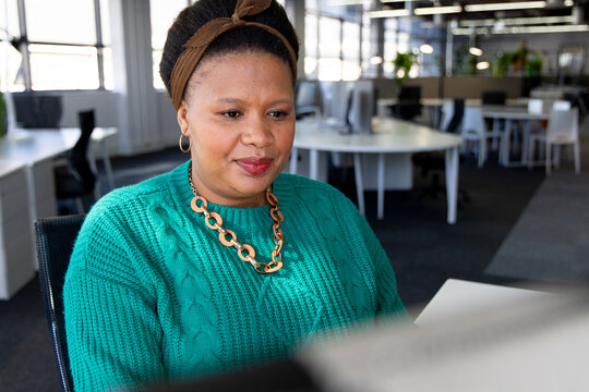 African American woman typing report on computer at desk in open-plan office with potted plants