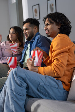 Diverse friends grasping popcorn boxes on sofa at home with abstract art reacting with disgust