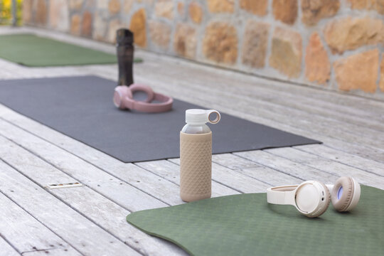Beige reusable water bottle is standing on wooden deck between two yoga mats, headphones nearby