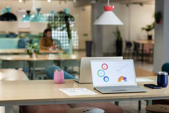 Open silver laptop is displaying charts and graphs on wooden table with pink and navy tumblers