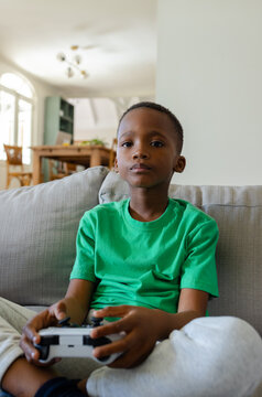 African American child in green tee sitting on gray sofa in living-room holding white black gamepad