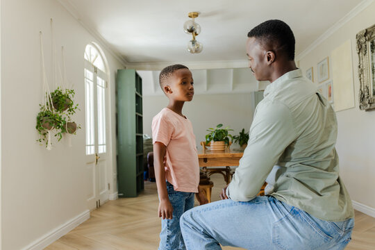 African American father kneeling, interacting with child boy in dining area near arched glass door