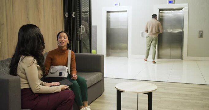 Diverse coworkers sitting office sofa using tablet noticing man pressing elevator button walking