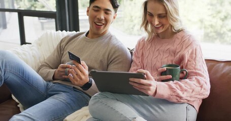 After holding credit card, diverse couple on couch tapping tablet making purchase sipping coffee - Powered by Adobe
