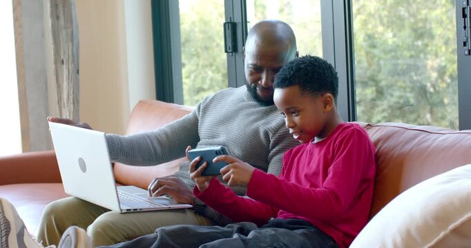 African American father and son playing smartphone game, father pausing typing, leaning in on couch