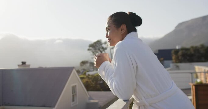 Sunrise drawing mid adult Asian woman onto balcony leaning on railing sipping mug, copy space