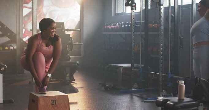 Woman stepping onto box tying purple shoes in gym, partner entering stretching on cable machine