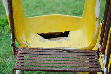Close-up of a weathered yellow playground slide showing chipped paint, a large hole in the seat area, and a rusted metal frame. Great for themes of decay, neglect, and worn outdoor playgrounds.